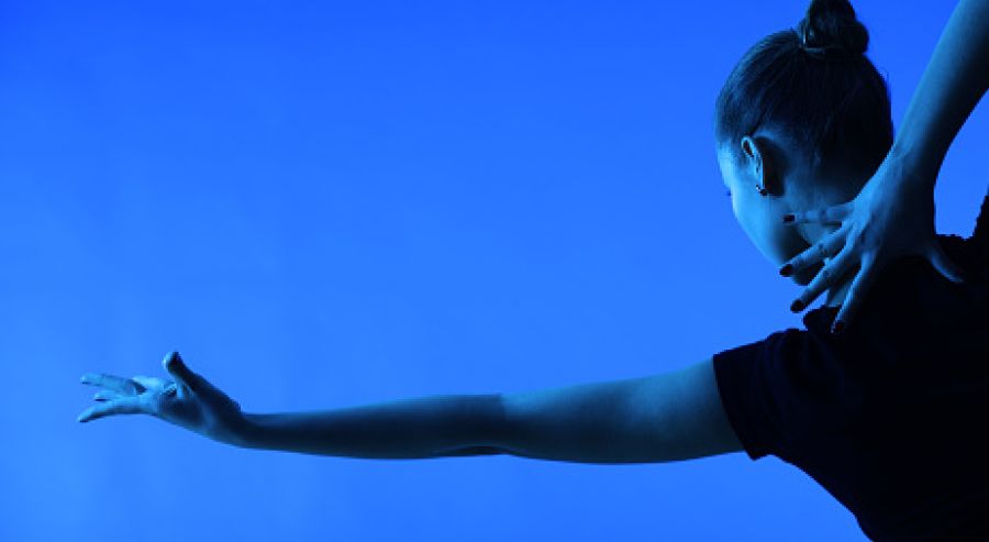 A person with their hair tied in a bun, viewed from behind, extending their left arm outward while placing their right hand on the back of their neck, set against a blue background.