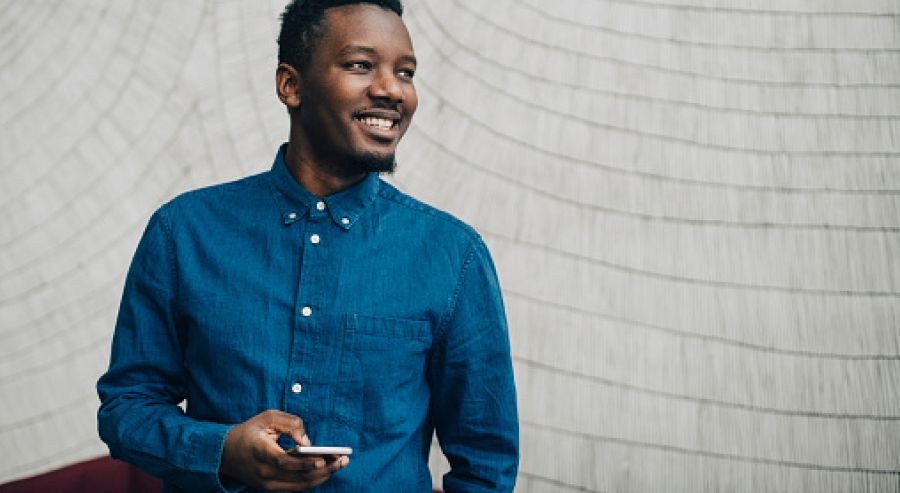 A person in a blue button-up shirt holding a smartphone in their right hand, standing against a light-colored wall with horizontal lines; the person's face is blurred.